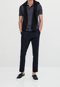 Navy polo with cable knit texture, black sweater casually draped over shoulders, dark pants, and black sneakers with white soles.