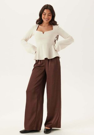 Kvinde smilende med langt mørkt hår, iført en elfenbensfarvet bluse med lange ærmer og brede brune bukser, stående med hænderne på hofterne.