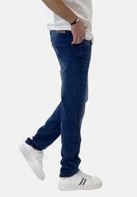 Jeans de mezclilla azul oscuro con corte ajustado, con bolsillos estándar y detalles de costura sutiles, combinados con zapatillas blancas. Camiseta blanca lisa.