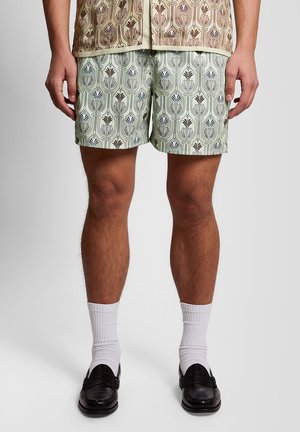 Man die een korte broek met bloemenmotieven draagt, gecombineerd met witte sokken en zwarte loafers, staand tegen een effen achtergrond.