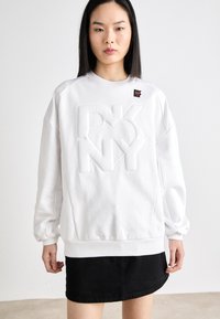Fehér pulóver domború "DKNY" logóval, bordázott mandzsettákkal és aljjal, kerek nyakkivágással, valamint fekete kiegészítő címkével a vállnál. Fekete szoknyával párosítva.