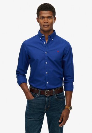 Blue long-sleeve button-down shirt with a collar, featuring white buttons and a small red logo on the chest, paired with dark jeans and a brown belt.