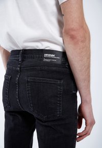 Jeans de mezclilla negra con un diseño ajustado. Presentan un parche con el logo en la parte trasera y dos bolsillos traseros, mostrando una textura suave.