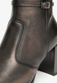 Bottine en cuir noir avec un talon carré, un panneau latéral, des détails de couture et une accentuation de boucle sur la sangle de la cheville.