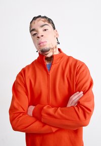 Sweatshirt vermelho com fecho zip, colar alto e mangas longas, feito de tecido suave, usado por uma pessoa com os braços cruzados.