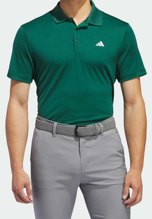 Mężczyzna w ciemnozielonej koszulce polo Adidas wpuszczonej w jasnoszare spodnie z szarym plecionym paskiem, stojący z luźno opuszczonymi ramionami.