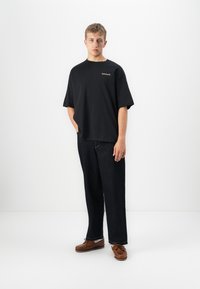 Un jeune homme se tient devant un fond blanc, portant un t-shirt noir oversized Timberland, un jean foncé et des chaussures bateau marron.