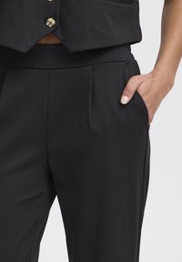 Black tailored pants with a smooth texture, featuring front pleats and side pockets. The waistband is elastic for comfort and fit.