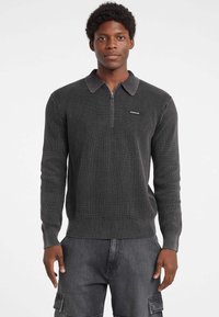 Halli halliga pullover, millel on tõmblukuga eesmine osa, krae ja pikad varrukad. Hallil pulloveril on rinna peal logo detail ja tekstuuriga kudum.