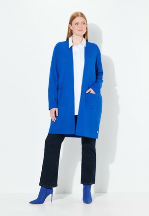 Lange blauwe cardigan met open voorkant, twee voorkuiste zakken en geribbeld textuur. Gecombineerd met een witte shirt en donkere jeans, met blauwe hoge hakken.