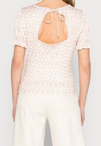 Top con estampado floral y mangas cortas con volantes, fondo de color rosa claro, espalda abierta con lazo, hecho de una tela suave y texturizada.