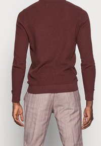 Persona con un maglione lungo a maniche lunghe di colore bordeaux e pantaloni a quadri rosa chiaro, mostrata di spalle con le braccia rilassate ai lati.