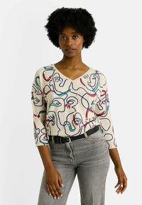 Pull beige à col en V avec un art linéaire abstrait multicolore, manches 3/4, associé à un jean gris taille haute et une ceinture noire.
