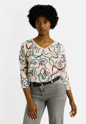 Suéter beige de cuello en V con arte lineal abstracto multicolor, mangas 3/4, combinado con jeans gris de cintura alta y un cinturón negro.