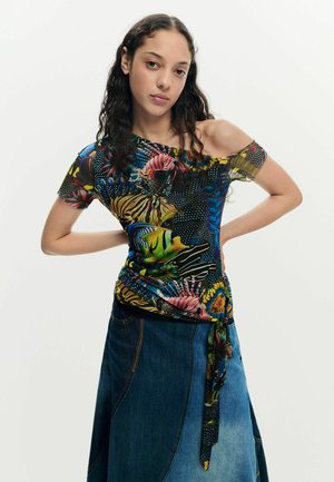 Noor naine pikkade lokkis juustega, kellel on seljast avatud troopiliste kaladega mustriga top ja kahetooniline teksaseelik, poseerib käed puusadel.