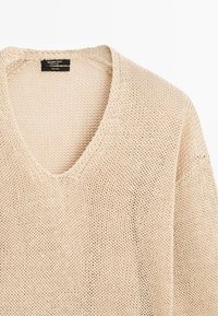 Beżowy sweter dziergany z dekoltem w kształcie litery V, charakteryzujący się luźną, otwartą teksturą. Wykonany w 100% z lnu, o luźnym kroju i opadających ramionach.