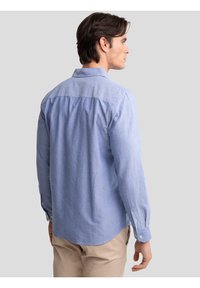 Bărbat văzut din spate, purtând o cămașă albastru deschis cu mâneci lungi și pantaloni bej, pe un fundal simplu gri deschis.