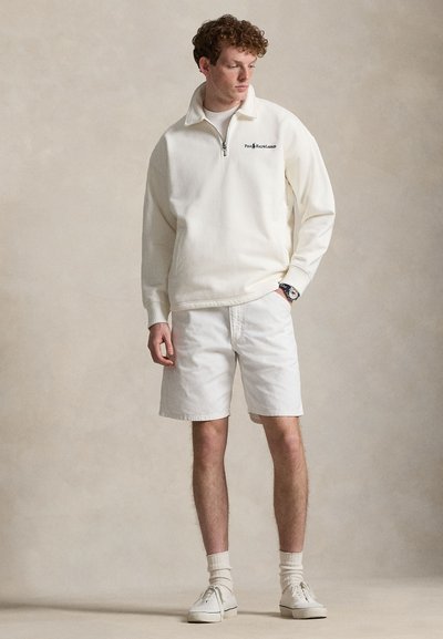 Νεαρός άνδρας με λευκό φούτερ με φερμουάρ μέχρι το στήθος, λευκά σορτς, εκρού κάλτσες και λευκά αθλητικά παπούτσια, στέκεται με το ένα χέρι στην τσέπη, μπροστά σε ουδέτερο φόντο.