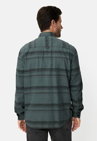 Dunkelgrünes Flanellhemd mit schwarzen und blauen Streifen, Knopfmanschetten und einer angenähten Schlaufe am Kragenrücken.