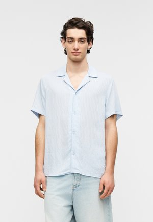 Hellblaues kurzärmeliges Hemd mit subtiler vertikaler Struktur, durchgehendem Knopfverschluss und offenem Kragen. Kombiniert mit hellen Jeanshosen.