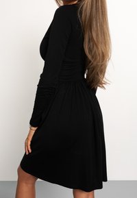 Robe noire à manches longues avec un corsage ajusté et une jupe évasée. Confectionnée dans un tissu lisse au toucher doux avec de subtils plis.