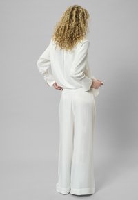 Weiße schillernde Bluse, kombiniert mit passenden weit geschnittenen Hosen, die einen subtilen Glanz aufweisen und lange Ärmel mit geknöpften Manschetten haben. Einfaches, elegantes Design.
