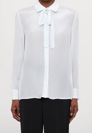 Kvinde iført en gennemsigtig hvid bluse med lange ærmer, frontknaplukning og bundet hals sløjfe, kombineret med sorte bukser.