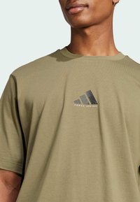 T-shirt in cotone verde oliva, con maniche corte e scollatura rotonda, caratterizzata da un logo a tre strisce nero sul petto.