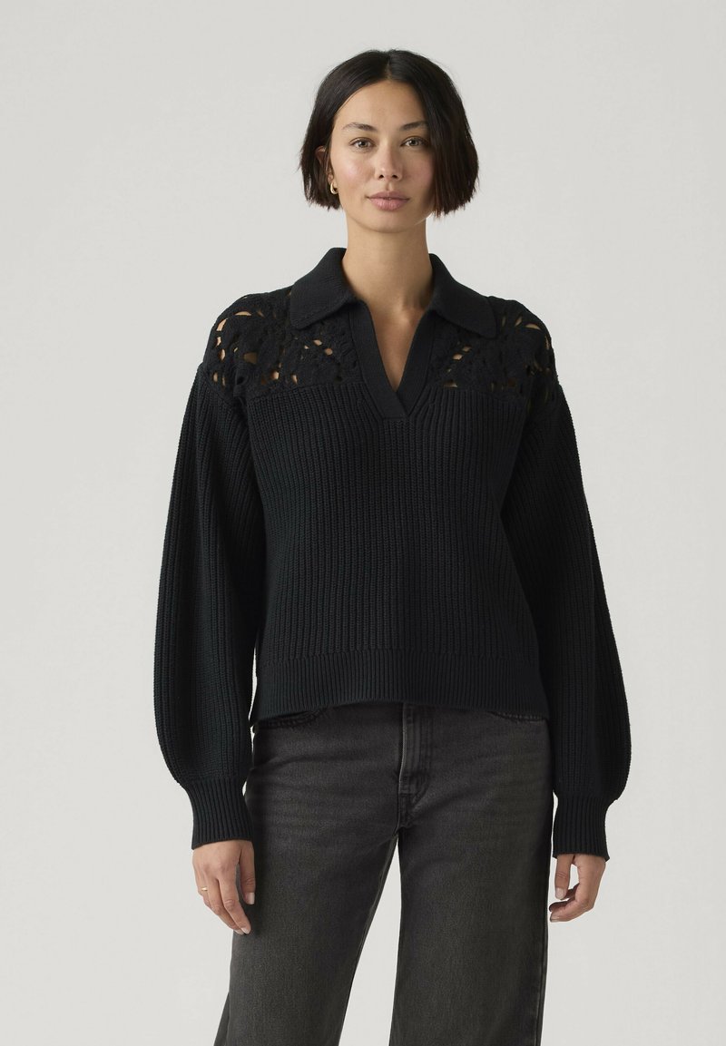Vrouw met kort donker haar, gekleed in een zwarte gebreide trui met kanten inzetstuk en kraag, gecombineerd met donkergrijze spijkerbroek, staand tegen een effen achtergrond.