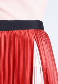 Falda con pliegues de acabado satinado rojo, que presenta una cinturilla elástica negra y un forro interior blanco visible a través de una apertura lateral.