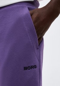 Σκούρο δέρμα στο χέρι που έχει τοποθετηθεί στην τσέπη μοβ παντελονιού με μαύρο κεντητό λογότυπο "BORG" κοντά στην τσέπη.