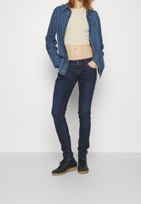 Denimskjorta över en beige crop top, tillsammans med mörkblå skinny jeans och svarta sneakers med gummisulor. Släta texturer och åtsittande design.