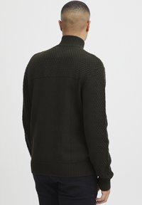 Dunkelgrüner Pullover mit hohem Kragen, der strukturierte Muster an den Schultern sowie gerippte Bündchen und Saum aufweist. Insgesamt glattes Strickgewebe.