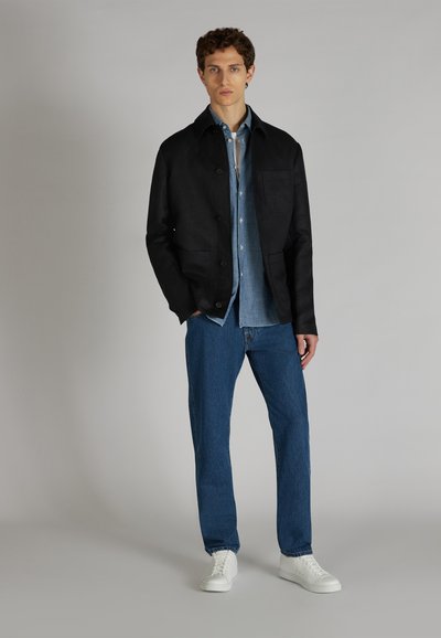 Giacca nera con bottoni sopra una camicia di denim, abbinata a jeans blu e sneakers bianche. Materiale morbido, vestibilità sartoriale, design minimalista.