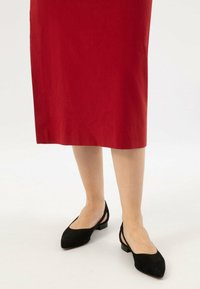 Nogi w czerwonej spódnicy midi i czarnych butach na obcasie typu slingback z niskim obcasem na białym tle.