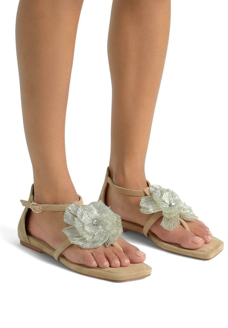Beige platte sandalen met een bloemenontwerp in zilver gaas op de teen, voorzien van een enkelbandje en een gladde, platte zool.