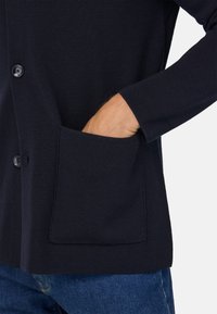 Jūras spēku krāsas adīta kardigāna žakete ar teksturētu virsmu, vienu pogu un kabatu. Redzama roka, kas sniedzas iekšā kabatā.