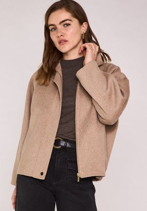 Femme aux cheveux châtain clair portant une veste beige zippée, un haut marron foncé, un jean noir et des boucles d'oreilles créoles dorées sur un fond uni.