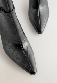 Bottes noires à la cheville en matériau synthétique brillant avec une texture crocodile, dotées d'un bout pointu et d'un design épuré.