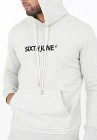 Sudadera con capucha gris claro y un bolsillo frontal, con "SIXTHJUNE" impreso en negro. Incluye cordones con puntas de plata. Diseño casual.