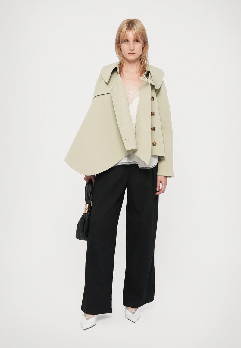 Veste cape vert clair avec un col et de grands boutons, portée sur un haut blanc et un pantalon large noir. Chaussures blanches pointues et sac noir.