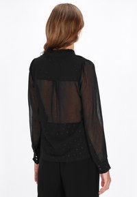 Blusa nera in tessuto semi-trasparente, colletto arricciato e dettagli scintillanti sottili. Maniche lunghe con polsini a bottone e una vestibilità rilassata.
