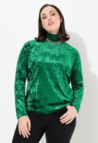 Top verde in velluto a maniche lunghe con collo alto, caratterizzato da una texture lucida e una vestibilità rilassata. Abbinato a pantaloni neri aderenti e gioielli in argento.
