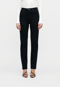 Γυναίκα με εφαρμοστά μαύρα τζιν, λευκό μακρυμάνικο τοπ και μαύρα ψηλοτάκουνα σανδάλια με λουράκια, που στέκεται μπροστά σε ένα απλό φόντο.