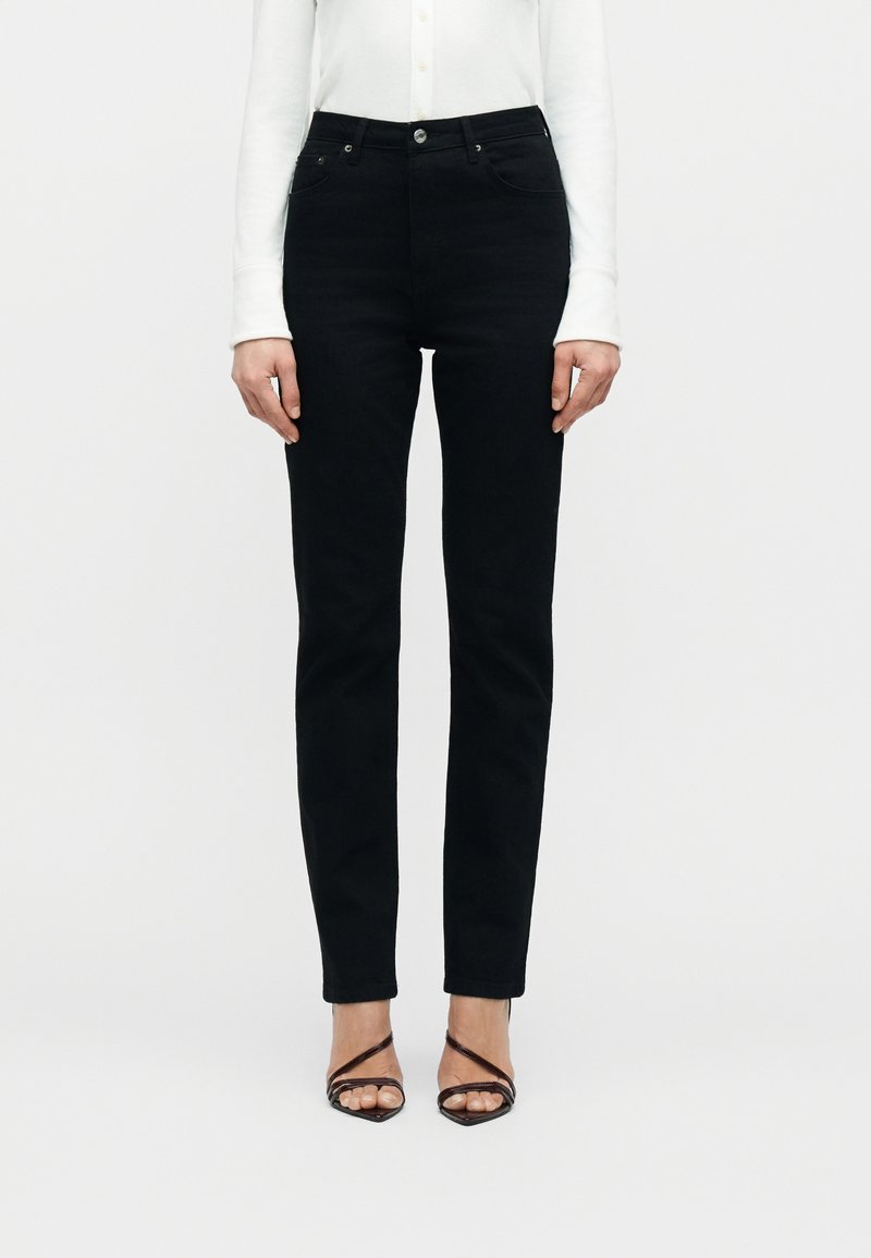 Γυναίκα με εφαρμοστά μαύρα τζιν, λευκό μακρυμάνικο τοπ και μαύρα ψηλοτάκουνα σανδάλια με λουράκια, που στέκεται μπροστά σε ένα απλό φόντο.