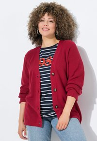 Roter, gerippter Cardigan aus strukturiertem Stoff, mit großen dunklen Knöpfen, über einem navy gestreiften T-Shirt getragen, kombiniert mit hellen Jeans.