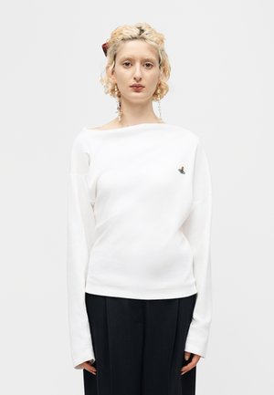 Ung person med krøllet blondt hår, iført perleøreringe, en hvid langærmet top med et lille emblem og sorte bukser, stående foran en ensfarvet baggrund.