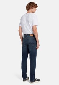 Jeans in denim blu scuro con un design a gamba dritta, dotati di un'etichetta in pelle beige, tasche posteriori con dettagli di cucitura, indossati con una maglietta bianca.