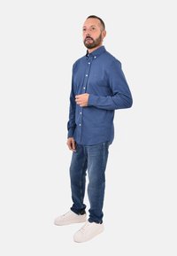 Camicia azzurra in cotone a maniche lunghe con colletto botton-down e bottoni frontali, abbinata a jeans in denim blu e sneakers bianchi.