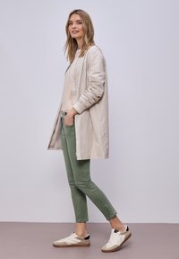 Giovane donna in piedi con una mano in tasca, indossa una giacca lunga beige chiaro, un maglione pallido, pantaloni verdi aderenti e sneakers bianche.